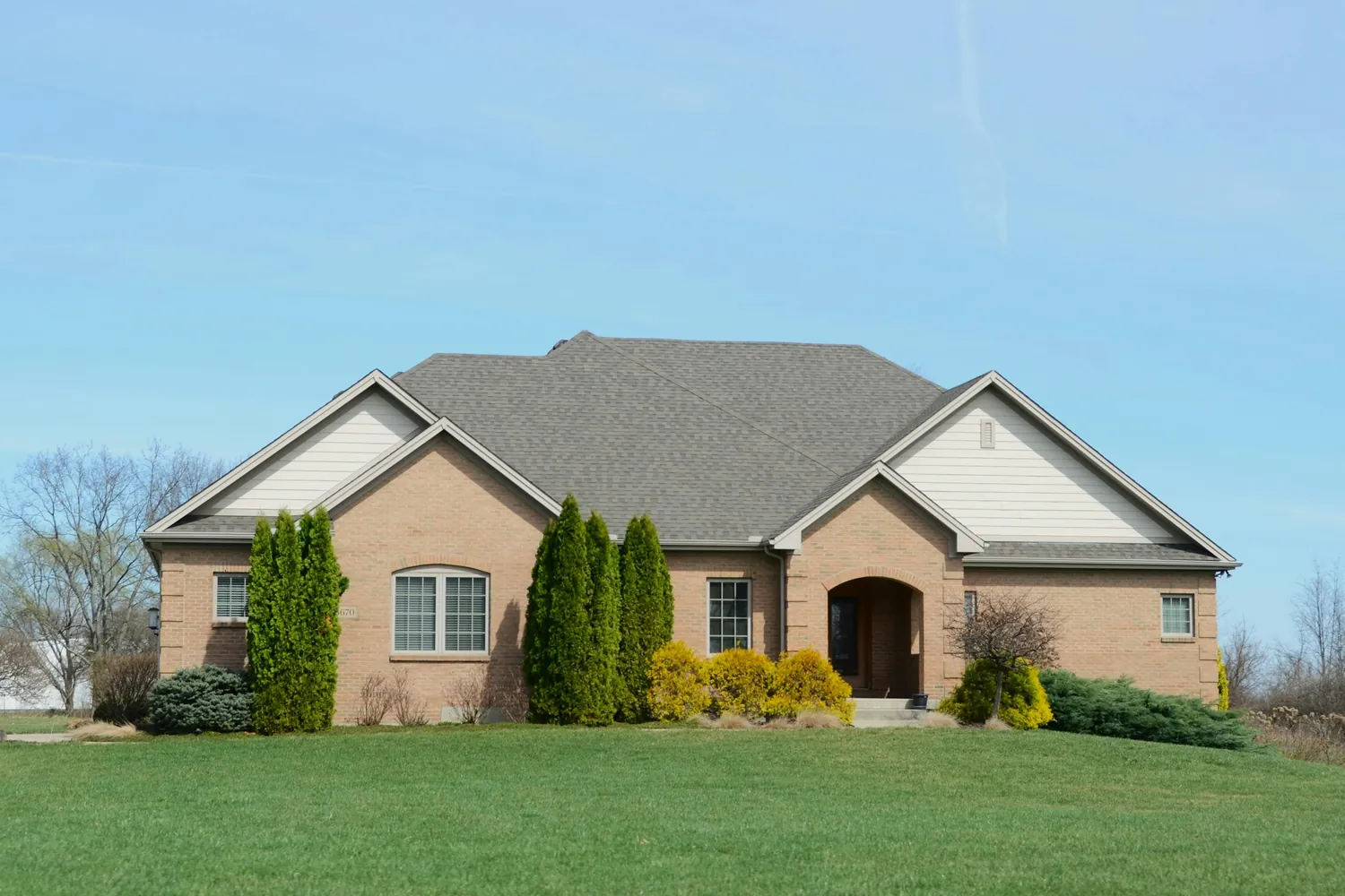 Tan brick single-story home with evergreen shrubs under a blue sky